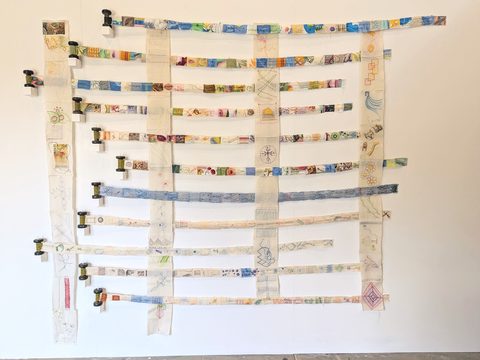 Woven grid of stitched calico strips suspended from cotton reels on a gallery wall